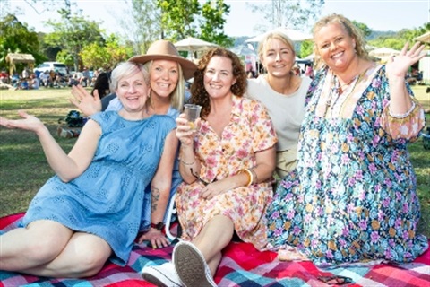 Five women sitting on a picnic blanket, one holding a cup