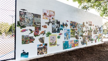 Photograph of a long white brick wall covered with installed collage artworks featuring animals, plants, landscapes.