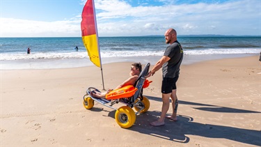 Person using a beach wheelchair on the sand near a surf lifesaving flag with a beach access assistant.