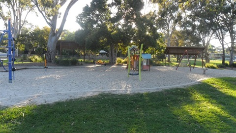 Apex Park - playground: the image shows play equipment set in a large sandpit sheltered by tall gum trees. There are two sheltered picnic tables in the background.