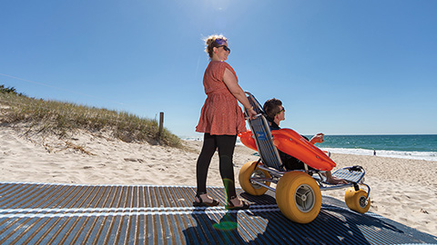 Image of a woman pushing a disabled person using beach accessible equipment.
