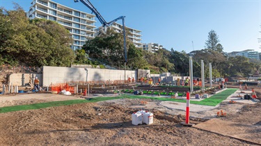 Concrete pump truck extending a long blue boom across an active construction site, where workers are preparing reinforced concrete foundations. Safety barriers, exposed steel reinforcement, and retaining walls are visible, with trees and multi-storey residential buildings in the background.