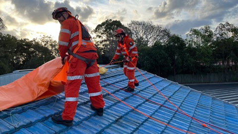 Two people wearing high vis safety gear, standing on a blue roof laying down a tarp.
