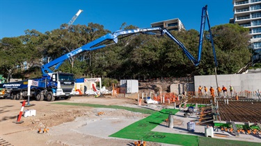 Wide view of a construction site showing reinforced concrete foundation works, vertical concrete columns, and workers within a fenced work area. A concrete pump boom extends above the site, with apartment buildings, trees, and clear blue skies in the background.