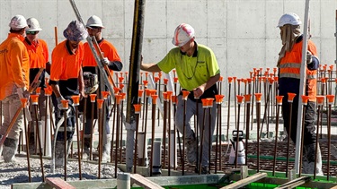 Construction workers placing and finishing concrete within reinforced foundation formwork. Steel reinforcement bars fitted with orange safety caps surround the work area, with a concrete retaining wall visible in the background.