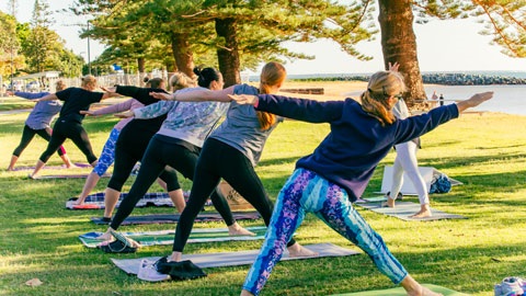 Group of people exercising outdoors on grass, stretching sideways in a yoga class in a park beside a river, with trees casting shade and buildings visible across the water.