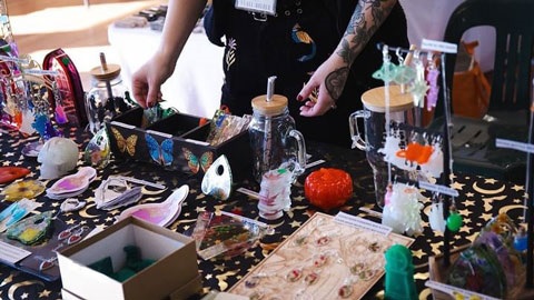 An arm reaches out to pick up something off a table that is covered in objects such as crystals and candles.
