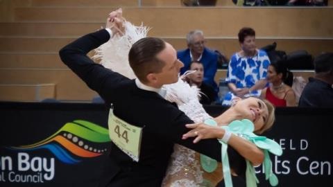 A man and women are dancing, the women is wearing a white ballroom dancing dress and the man is in a tuxedo. 