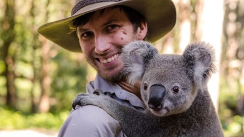 A man in khaki is looking at the camera smiling while holding a koala.