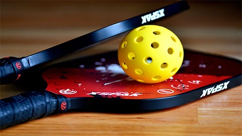 A pickleball ball is resting between two pickleball rackets on the wooden floor.
