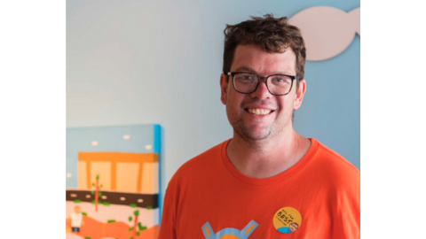 A man in glasses wearing an orange shirt smilling at the camera.