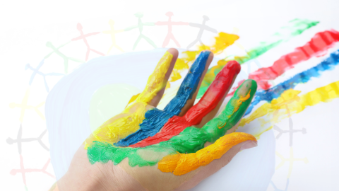 A hand held palm up with yellow, blue, red, green and orange paint on each finger. In the background, is a circle with stick inger people around it.