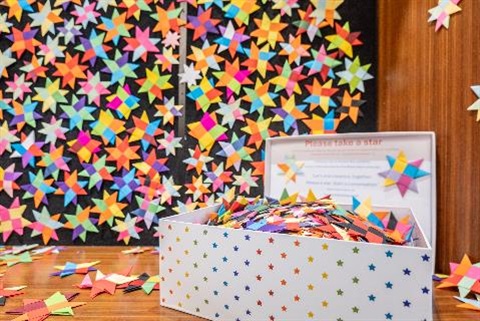 Multi-coloured stars line the entire backdrop, they are made from paper, by victims of DFV. In the forground, is an open wooden chest, decorated in shiny stars.