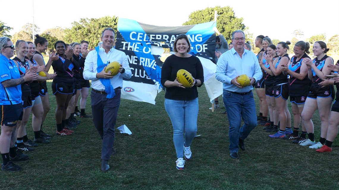 AFL Queensland manager Damien Mitchelmore, Pine Rivers MP Nikki Boyd and City of Moreton Bay Mayor Peter Flannery run through a banner while Pine Rivers Swans players cheer them on