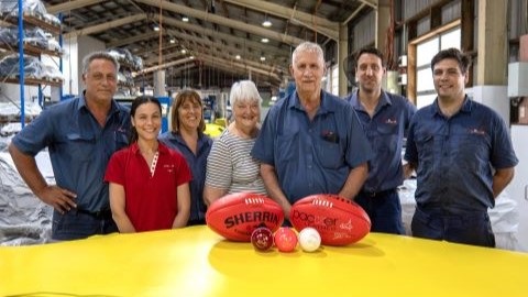 The Packer Family members who work at Packer Leather. The family members are standing in the factory smiling with a stack of yellow leather in front of them with some cricket balls and two AFL balls on it.