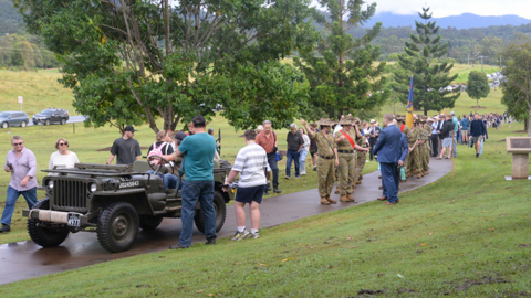 An old army truck on a road in Samford with locals watching and service men and women parading behind it. There is green grass and mountains rolling behind it.