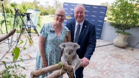Moreton Bay Wildlife Hospital Foundation Director Christine West and City of Moreton Bay Mayor Peter Flannery are standing behind a koala on a tree branch. They are both smiling and the koala is posing in front of them among tree branches and gum leaves.