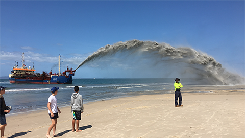 Beach nourishment work in action, during a previous campaign. A ship shooting sand onto a beach can be seen in the distance, being watched by onlookers.