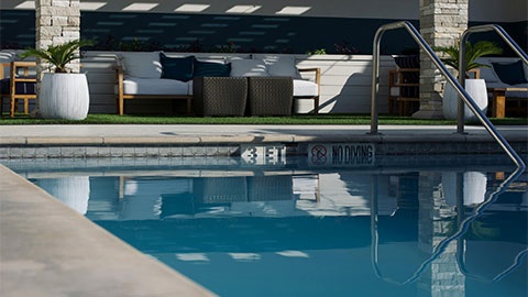 In the background there's outdoor seating on grass with a potted plants surrounding the seats. A pool is in the foreground with a safety rail going into the pool.