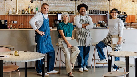 Two women and two men are facing towards the camera smiling. The women are half sat on high stools and the men are leant against a high bar. In the background there is a wall of spirits, liquor, stemmed glasses and cups. In the foreground there are dining tables and chairs pushed in.