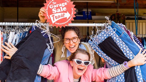 2 women smiling pushing through a rack of second-hand clothes and holding a small red sign 'The Garage Sale Trail'.