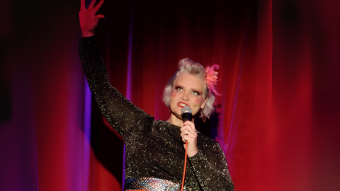 a woman with blonde hair and stage makeup standing in front of a stage curtain with a microphone singing