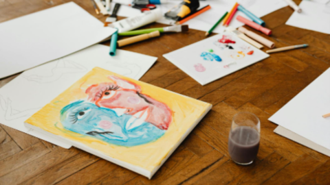 An abstract portrait is on a wooden table, there are art supllies scattered behind it,