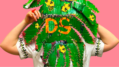 Photograph of a person wearing a green leaf‑like sculptural mask with yellow floral details against a pink background.