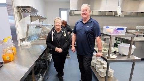 Michelle Gilchrist and Mayor Peter Flannery standing in the brand ne kitchen for The Breakfast Club at the Peninsula Support Hub opening. To their left is orange juice and behind them is the breakfast food ready to be served. The kitchen is full of bench space and large appliances. Michelle and Mayor Flannery have a big smile on their faces happy to see the facility open and meal services running