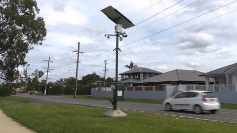 A car travelling on a road with a CCTV camera set up beside it. There is a green nature strip and then a pathway runnign alongside the road. Ther are powerlines and lights alongside the road as well. 