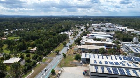 A street cuts diagonally through the image. On the left is green space with houses dotted in between, and on the right are businesses. There is blue sky and clouds in the distance and above the street.