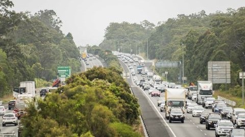 Heavy traffic on a multi‑lane highway with cars and trucks travelling in both directions, bordered by dense bushland and roadside signage.