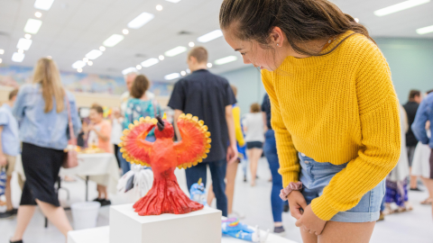 Person viewing a colourful handmade artwork displayed on a pedestal at a community art exhibition, with visitors and exhibits in the background.