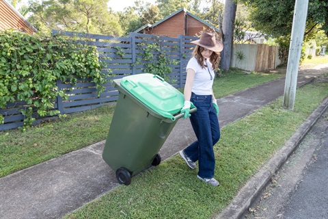 Person wheeling a green rubbish bin along a suburban footpath beside a grass verge and timber fences.