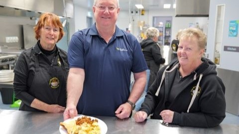 Mayor Peter Flannery is with Michelle Gilchrist and a volunteer from The Breakfast Club. They are smiling and standing in the kitchen holding out a plate with warm breakfast on it..jpg