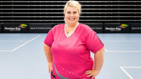 Person standing on an indoor sports court, wearing a pink sports top, with hands on hips and seating visible in the background.