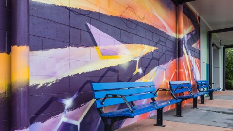 a mural painted on a large wall behind public seating with purple orange yellow white and green colours forming a mural for the community to enjoy