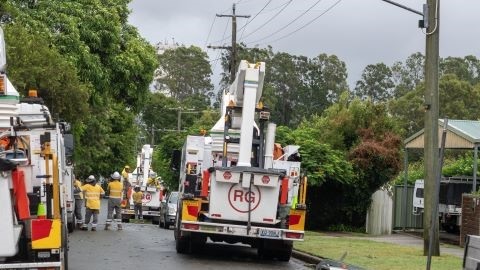 Energex trucks and workers lining a street with fallen trees and storm debris across the road. 