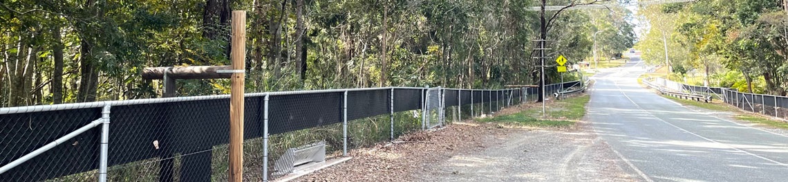 A fauna fence runs alongside a road. There is a wooden pole that goes over the fence where wildlife can climb over.