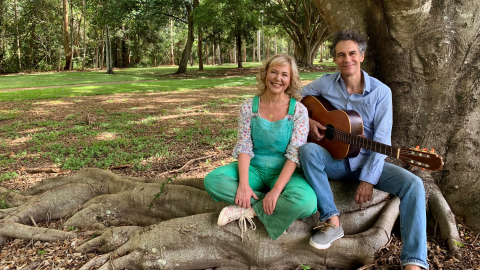 Two people sitting on a tree trunk holding a brown guitar and smiling