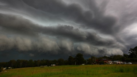 A grassy field under dark storm clouds. Trees and buildings can be seen in the distance. 