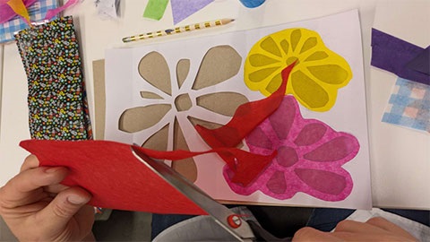 There's a table full of arts and crafts, in focus is a pieceof paper with flowers cut out and tissue paper glued over the top of the cut out flowers to provide them with colour. A person is using a pair of scissors to cut red tissue paper.