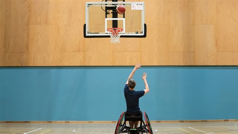 A person in a wheelchair, back facing the camera, is throwing a ball into a basketball hoop.