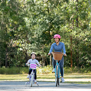 A mother, Emma, and her daughter, Lyla, are riding their bikes with their helmets on smiling at the camera.