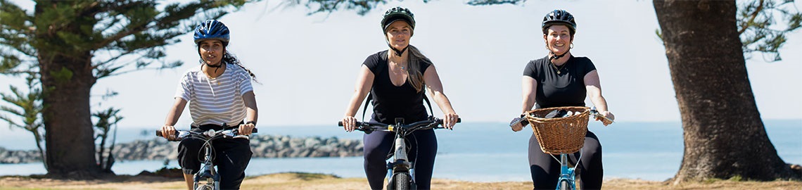 Three women are riding their bikes toward the camera. In the background is a sea front with trees on either side and a clear view over the water.
