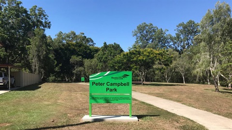 A green park sign reading 'Peter Campbell Park'. There are trees in the distance and a cement footpath to the right hand side.