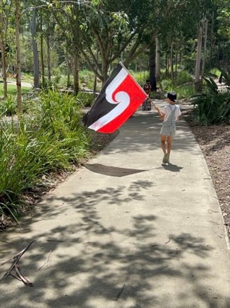 A child holding the national Māori flag, walking along the footpath surrounding by bushes and trees.