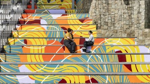 In the centre of the frame sit two people in conversation. They are sitting on stairs that fill the whole frame. The stairs are painted in yellow, oranges and dark reds, in an abstract pattern like a snake