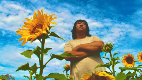 The camera is angled low looking up at a blue sky with some white clouds. In the forgeround is a field of sunflowers. Standing amongst the flowers is Rexy, a dark skinned man wearing glasses and a cream shirt. His arms are crossed.