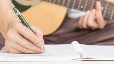 Image of a hand holding a pen and writing in a notebook while holding a guitar.